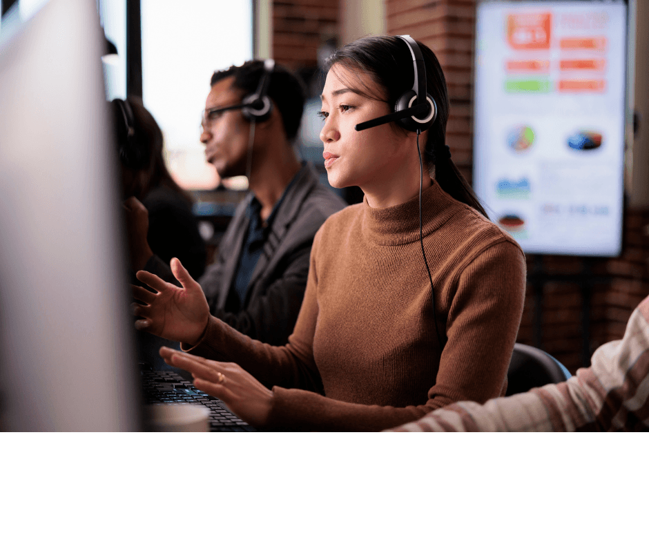 A professional engaged in a system audit, speaking into a headset while working on a computer in a collaborative office environment, with colleagues visible in the background.
